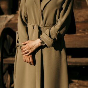 A stylish woman stands outdoors wearing a khaki trench coat and wristwatch, capturing fashion in a rustic setting.